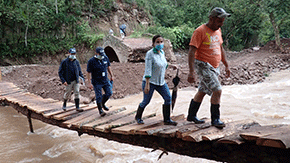 Two Project HOPE-sponsored health workers wearing navy blue shirts are being led by two people in orange and light blue as they all cross a rickety wooden bridge in Ecuador nearly  washed out by a hurricane.