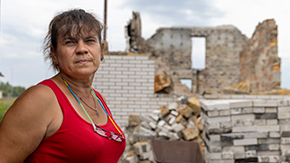 A woman with medium brown hair in a bun, wears a red tank top, silver necklace and glasses with on a strap around her neck. She stands in front of the ruins of a brick building under a cloudy sky.