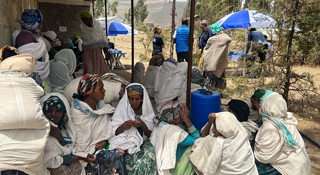 An outdoor scene in Ethiopia shows men and women in white linen body wraps over their brightly patterned clothing, sitting outside a building while a Project HOPE team in the background wears navy shirts and vests, setting up supplies and stations to provide medical care and consultations.