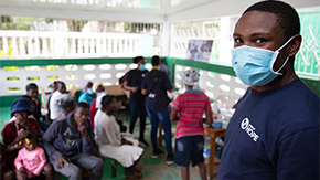 A Project HOPE-sponsored health worker is wearing a navy blue Project HOPE shirt and a blue medical mask as he stands in front of a group of people sitting in a clinic waiting room in Haiti. He is looking at the camera.