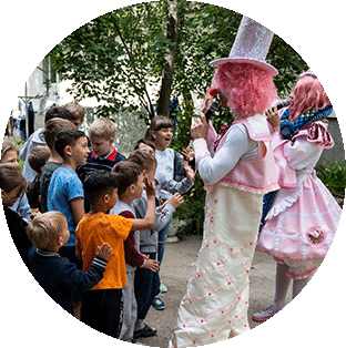 Project HOPE supports programming for Ukrainian children pictured here. Two clowns in white and pink costumes play pipe insturments for laughing and clapping children against a background of trees and white buildings.