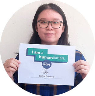 A round photograph of a  woman holding a 'I am a humanitarian' sign. She has long black hair and glasses. She is looking at the camera with a slight smile.