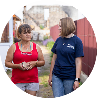 A photograph of two women wakling down an alleyway in Ukraine. One is a Project HOPE team member wearing a staff shirt. The woman she is talking to and looking at holds her phone in her hand and wears her glasses around her neck as she looks down pensively.