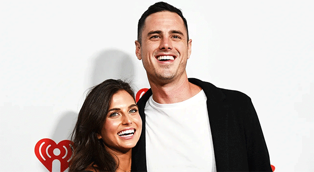 A photograph of The Bachelor stars Ben Higgings and Jessica Clarke against a white background at a press event. Both have big smiles and crinkled eyes. Ben wears a white tshirt and black sports jacket.