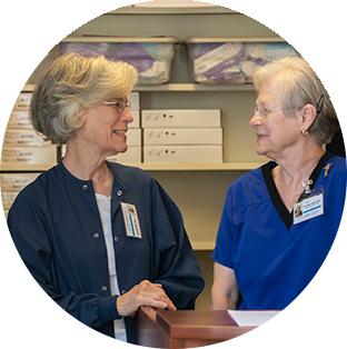 A round photograph of a two older woman with short gray hair looking directly at each other and smiling. The backdrop is a clinic with a wooden desk and medical supplies on shelves.