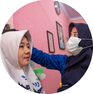 A round photograph of two women in Indonesia. One is a Project HOPE-supported health worker all in navy who is measuring the height of the other who wear a cream head scarf with blue floral decorations.