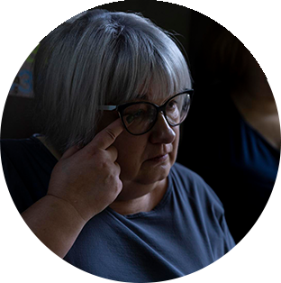 A Project HOPE-supported health worker in Ukraine with short gray hair and bangs wipes a tear from her eye with her finger. She wears black cat eye glasses and a sad expression.