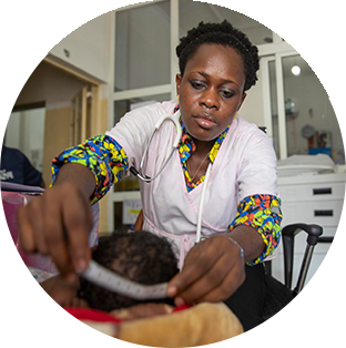 A photograph of a Project HOPE supported doctor in Sierra Leone looks intently as she wears brightly patterned clothers under her pink lab coat and measuring a baby with dark curly and thick hair.