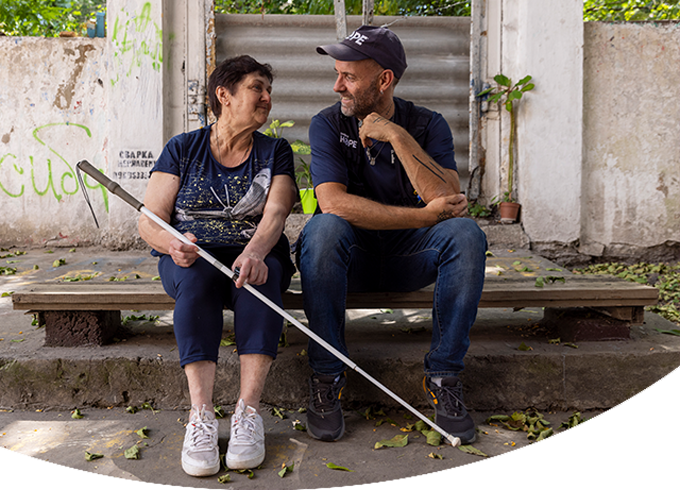 A member of the Project HOPE Emergency Response Team in Ukraine sits with an older women and beneficiary on steps outside. They are smiling at each other. Both wear navy blue shirts. The woman is holding a long silver cane.