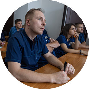 A photograph of a class of Ukrainian health workers. The main subject is looking pensively up to the right. They all wear navy blue scrubs and polo shirts as they sit and listen.