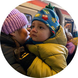 A Ukranian child is held by their mother as they wear a green parka, black gloves and blue hat. A train and crowd of people are in the background.