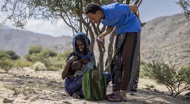 A mother holds her baby's ear open while a Project HOPE-supported health worker looks down at the child before giving them medicine. They stand outside against a dusty backdrop and scattered trees and shrubbery in Ethiopia.