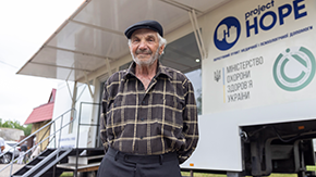 A photo of an older man in a cap smling as he stands in front of a Project-HOPE supported mobile clinic in Ukraine.