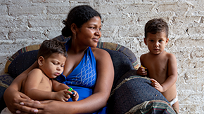 A refugee from Venezuela, a young pregnant woman in a halter top, sits on an armchair with one of her small children on her lap and the other looking at the camera shly at her side.