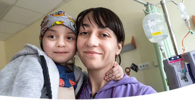 A mother and child from Ukraine stand against the background of an off-white hospital room in Poland. The mother holds her daughter in her arms as she wears a sweatshirt and her dark hair with bangs in a ponytail. The girl also wears a sweatshirt and gray cap as she hugs her mom with her arm around her neck.