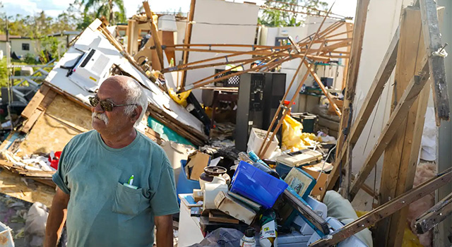 A photograph of an older man wearing a tshirt and tinted glasses as he is looking to the right and standing against the backdrop of the wreckage of his house after Hurricane Ida in 2022.