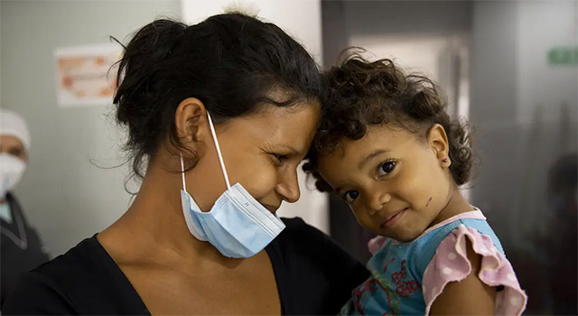 In Colombia, a mother wearing a black shirt and blue medical mask looks at her young toddler lovingly as she holds her in her arms. The toddler smiles at the camera and has curly dark hair and is wearing a blue and pink shirt.