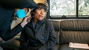 A women in Florida with dark hair and bangs with a dark jacket sits on a gray couch and looks to the right as she is being examined by a health worker in a blue medical mask.