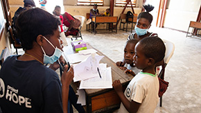 A Project HOPE-sponsored health worker is wearing a navy blue Project HOPE shirt and medical mask a d black pants as she stands sits at a wooden deakd and speaks to a family of two children and their mother in a clinic in Haiti.