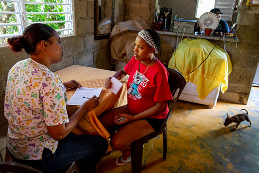 A photo of Yerika and a Project HOPE health worker in her home in the Dominican Republic. They sit at a table covered with a table cloth as they talk intently against the backdrop of a cinderblock room with a big window and a little dog walking by.