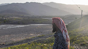 A photo of a man in Ethiopia named Abdul as he stands by a dry river bed showing the devastating impact of the drought there. He wears a patterned head covering and long-sleeve shirt as he looks out at the brown and green landscape.