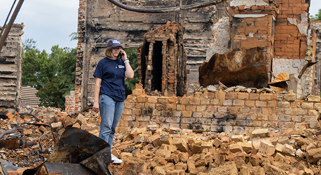 A photograph of a young person in a Project HOPE hat and tshirt walks through piles of rubble and twisted metal pieces as she looks down and talks on the phone. Pictured is Julia, Project Hope's Program Coordinator for rehablitation projects in Ukraine.