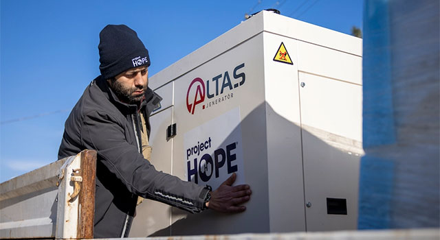 A photograph of a our local team member, Yunus, in a Project HOPE knit hat applying a sticker to a large square machine, a generator we delivered to Araban, Gaziantep province via the Turkish Ministry of Health.