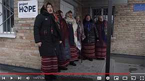 A screenshot of a paused video showing six women bundled in jackets, head scarves and skirts singing in front a brick building with a Project HOPE sign on the front. Pictured is a village clinic Project HOPE helped rebuild and stock after the Russian invasion.