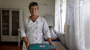 A photograph of a Ukranian woman with short hair, wearing a white nurse's uniform and holding onto a green first aid kit as it sits on a hospital cot. She has a serious and sad expression as she looks directly at the camera against the backdrop of a village clinic with lace curtains and large supply cabinet.