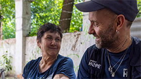 A photo of a Project HOPE staff member talking and smiling at an older Ukranian woman who smiles back at him, looking brightly at his face, against the backdrop of a concrete and barbed wire wall.