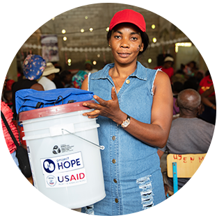 A round photograph of a woman in Haiti, wearing a cap, denim dress and watch, as she holds a water purifying bucket from Project HOPE.