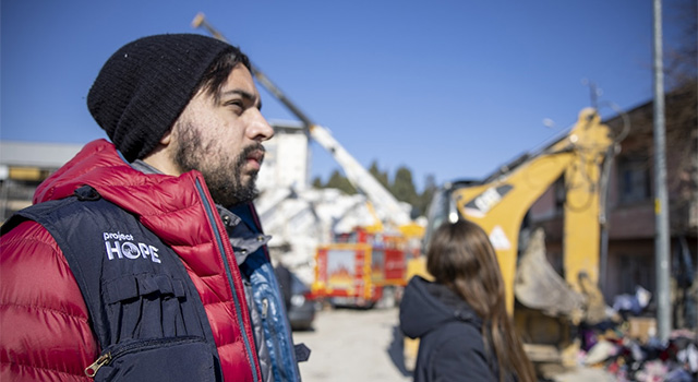 A photograph of a man's profile in a Project HOPE vest and red parka looking to the right. He is against a background of construction and rubble in Turkey.