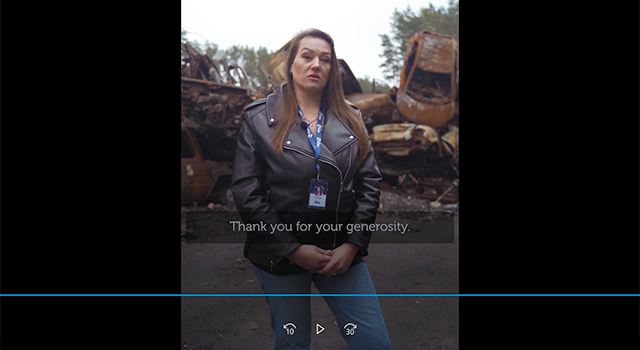 A screenshot of a paused video showing Vira, a Project HOPE team member, in front of a pile of burned cars. She is wearing a leather jacket and jeans and has long light brown hair.