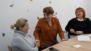 A photograph of three women in Ukraine wearing long-sleeved shirts. They are laughing and talking to each other as they sit at a wooden desk, covered with papers and pens, in wooden chairs, against the backdrop of a wall covered in stickers.