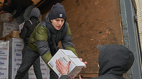 A photograph of a Project HOPE delivery in Ukraine of boxes labeled, Family Kit. A young person in a truck and heavy jacket and knit hat hands a box down to a waiting man.