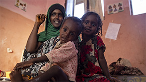 A photograph of a family in Ethiopia wearing brightly colored and patterened clothing. The mother and her two small children smile at the camera as they sit in a village clinic Project HOPE helped rebuild and stock.