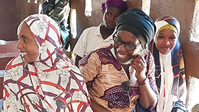 A photo of four women in Nigeria laughing as they sit together in a Project HOPE-supported health center. They wear brightly-colored and patterened clothing and headscarves against the backdrop of a brown wall with many windows.