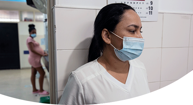 A photograph of Diana, the head nurse at a Project HOPE-supported hospital in Colombia. She is standing against a tile wall and wearing white scrubs and a blue face mask as a pregnant women shows blurrily in the background of a hospital room.