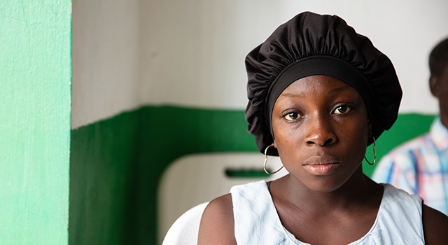 A photograph of a young woman wearing a tanktop, bonnet and heart-shaped earrings as she looks at the camera intently. She is sitting in a chair against the background of sparse exam room in Haiti.