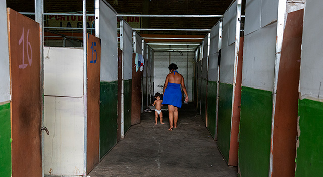 A photograph of the hallway of a temporary shelter for Venezuelans who have migrated to Colombia. We see a woman in a dress walking away with her back to the camera as she holds her young son's hand.