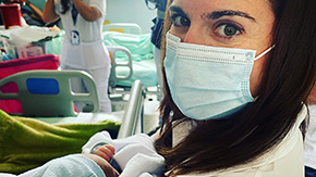 A photograph of a Project HOPE supported health worker in Colombia who is looking at the camera while wearing a medical face mask and holding a small baby who wears a hat and blanket.