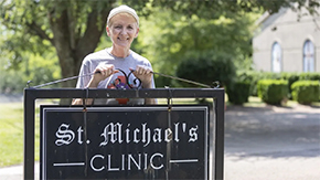 A photograph of a woman standing behind a large metal sign reading St. Michael's Clinic. she smiles at the camera against a sunny background outside.