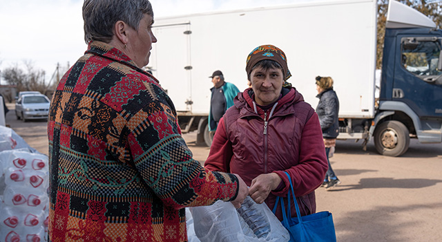 A photograph of two women as one hands the other a bag with 2 large bottles of water in the Kherson region of Ukraine. Both wear jackets and people walking among trucks and cars make up the background.