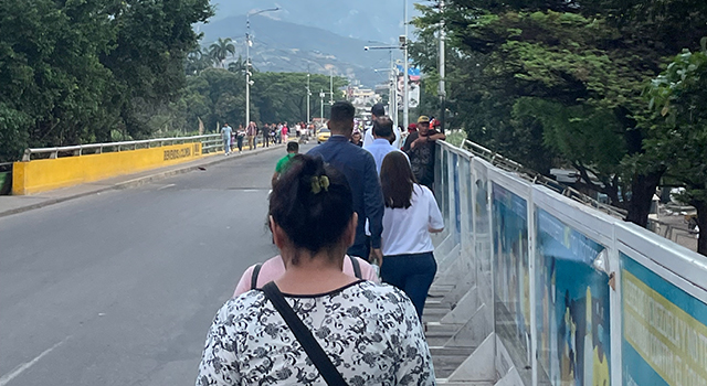 A photograph of a line of people people walking single-file down a sidewalk towards the border between Colombia and Venezuela. Trees, guardrails and lamposts line the empty roadway.