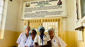 A photograph of 4 health workers smiling at the camera in front of a gated door to the National School of Midwifery in Sierra Leone. They all wear scrubs, lanyards and medical hair coverings.