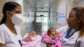 A photograph of two Project HOPE supported health workers in Colombia as they face each other in a hospital hallway. Both wear tshirts and holding a small baby, twins, in pink fuzzy blankets.