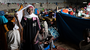 A photograph of a woman in a head scarf and track suit standing inside a displaced persons camp with corrugated metal walls. Tents, clothing and other supplies are scattered in the background where other families are living.