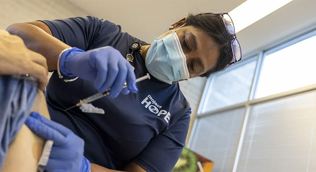 A photograph of a doctor in a Project HOPE tshirt, medical mask and gloves as she adminsters a shot to a patient. She is looking intently down with her glasses perched on her head and a stethoscope around her neck.