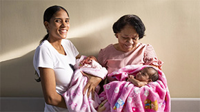 A photograph of a mother and health worker smiling at the camera as they each hold twin babies in a Project HOPE-supported clinic in Colombia.