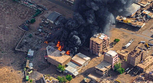 A photograph of burning buildings in Sudan where violence has broken out. Thick gray and black smoke makes up a large plume against a sandy backdrop.